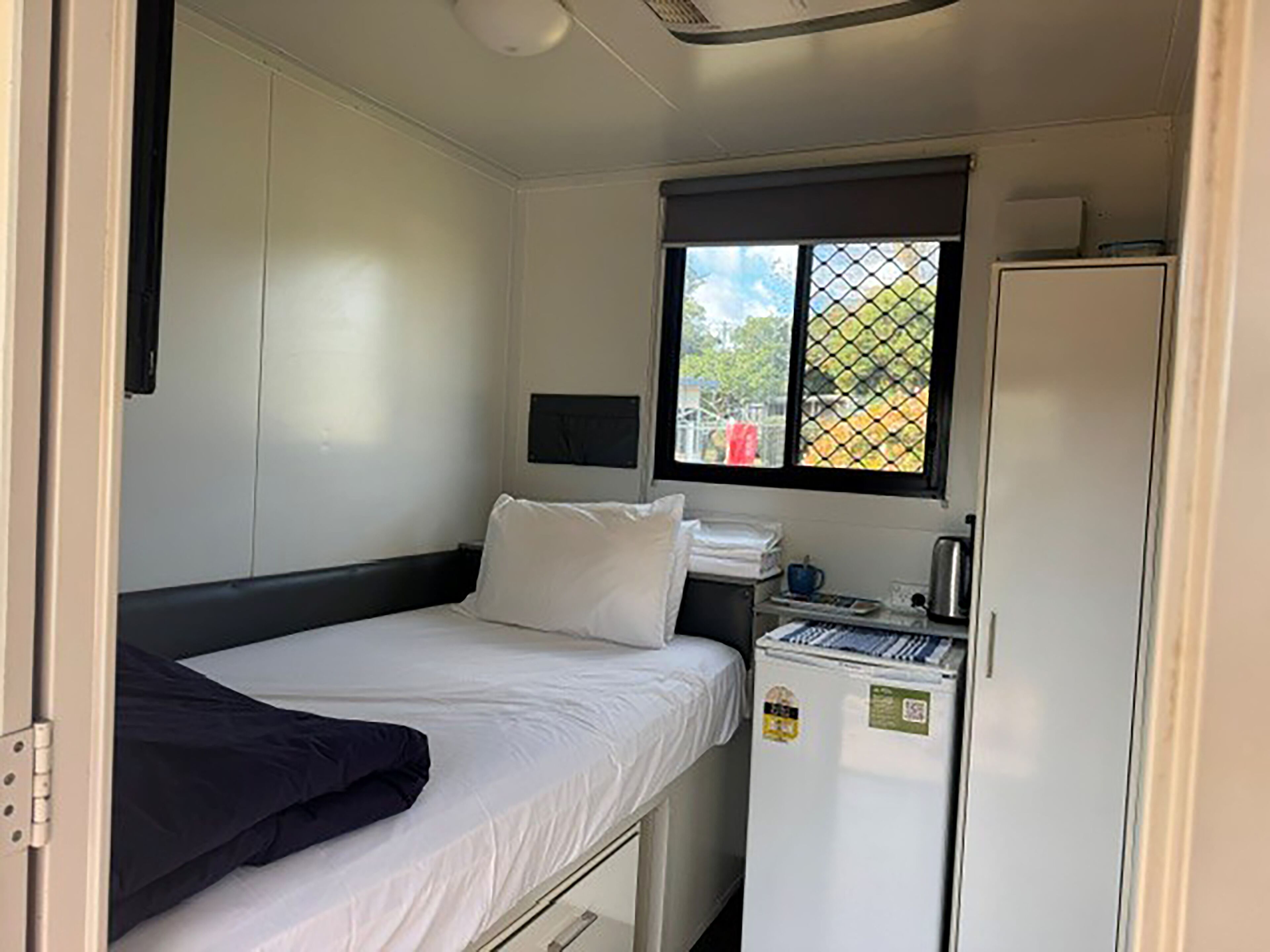 A small, single-bed room with a window, a small kitchenette, and a white wardrobe.