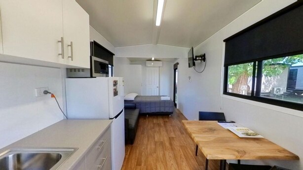 A compact kitchenette with a sink, a microwave, and a refrigerator. There is a bed with a gray mattress and a white pillow. A wooden table is positioned near a window with a view of greenery outside. - Camden Holiday Park (Elderslie)