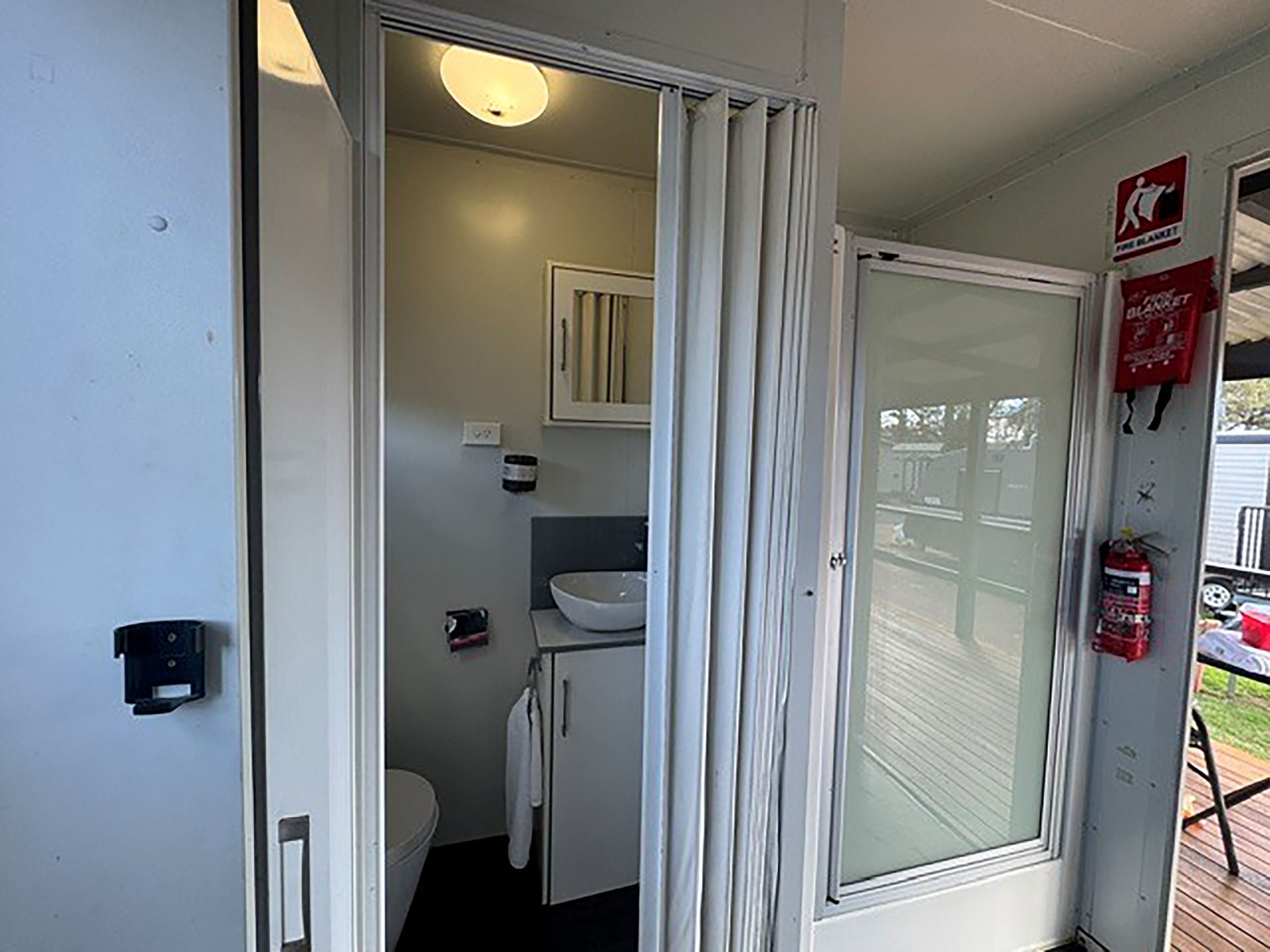 A bathroom with a toilet, sink, and cabinet, a glass door leading to an outdoor area, and a fire extinguisher mounted on the wall.