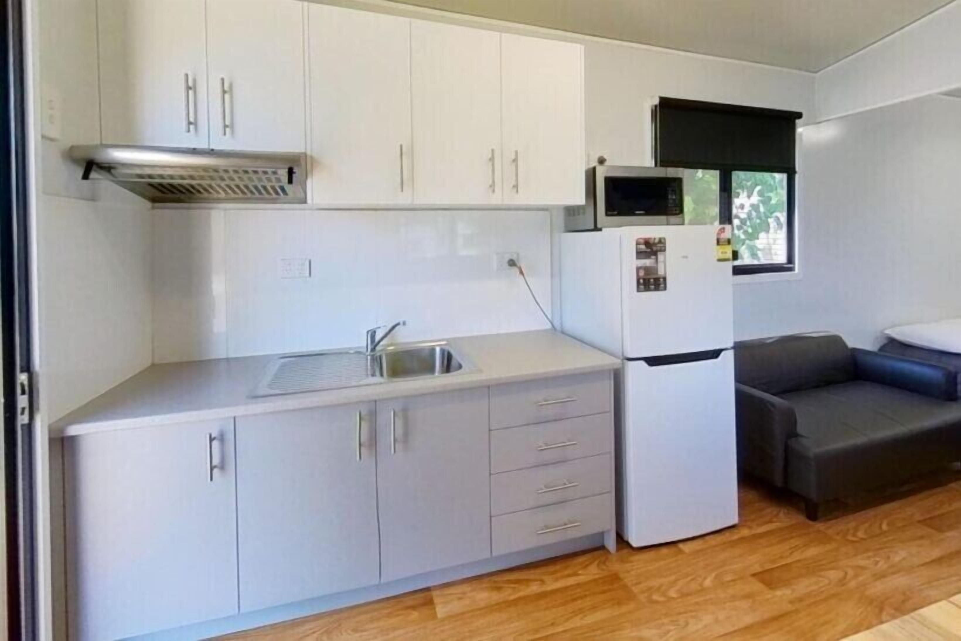 A compact kitchen with white cabinets, a sink, and a microwave. There is a dark sofa and a window with blinds.