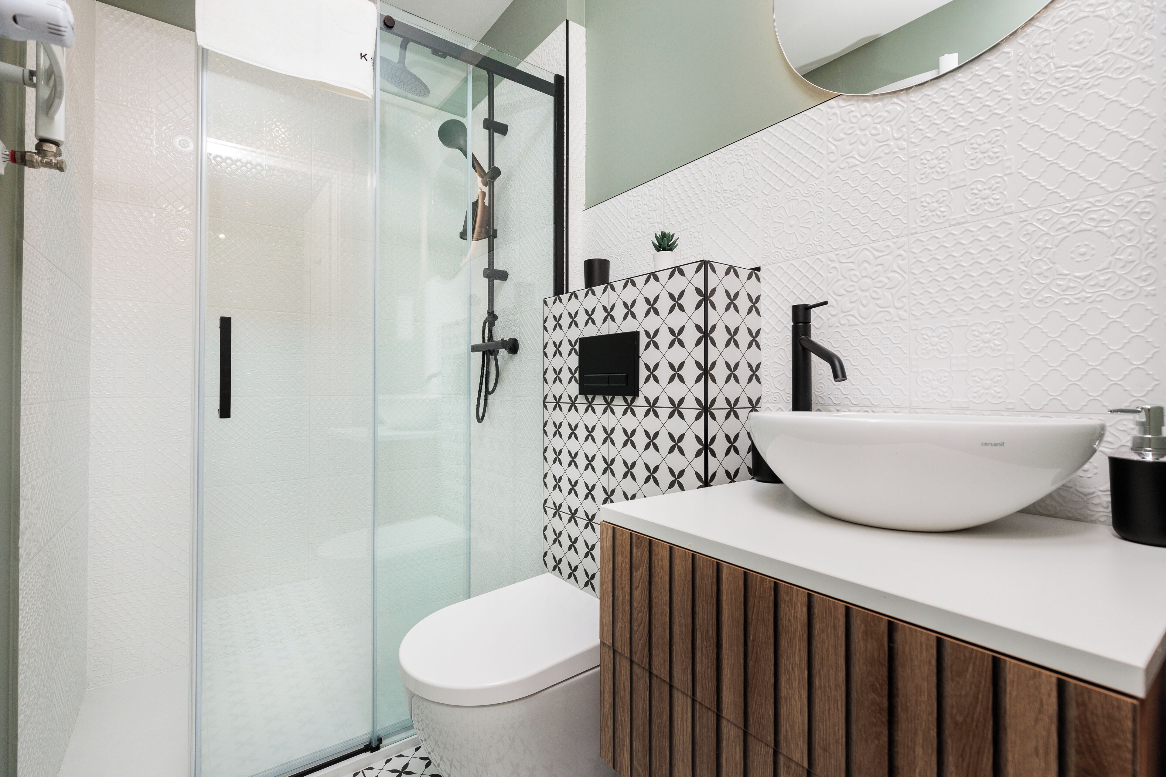 Ein modernes Badezimmer mit Duschkabine aus Glas, einer weißen Toilette, einem hölzernen Waschtisch mit weißem Waschbecken und einer gemusterten Spritzschutzeinrichtung.