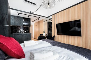 Quarto de hotel moderno com uma cama grande, televisão fixada na parede, escrivaninha com cadeira e área de kitchenette.