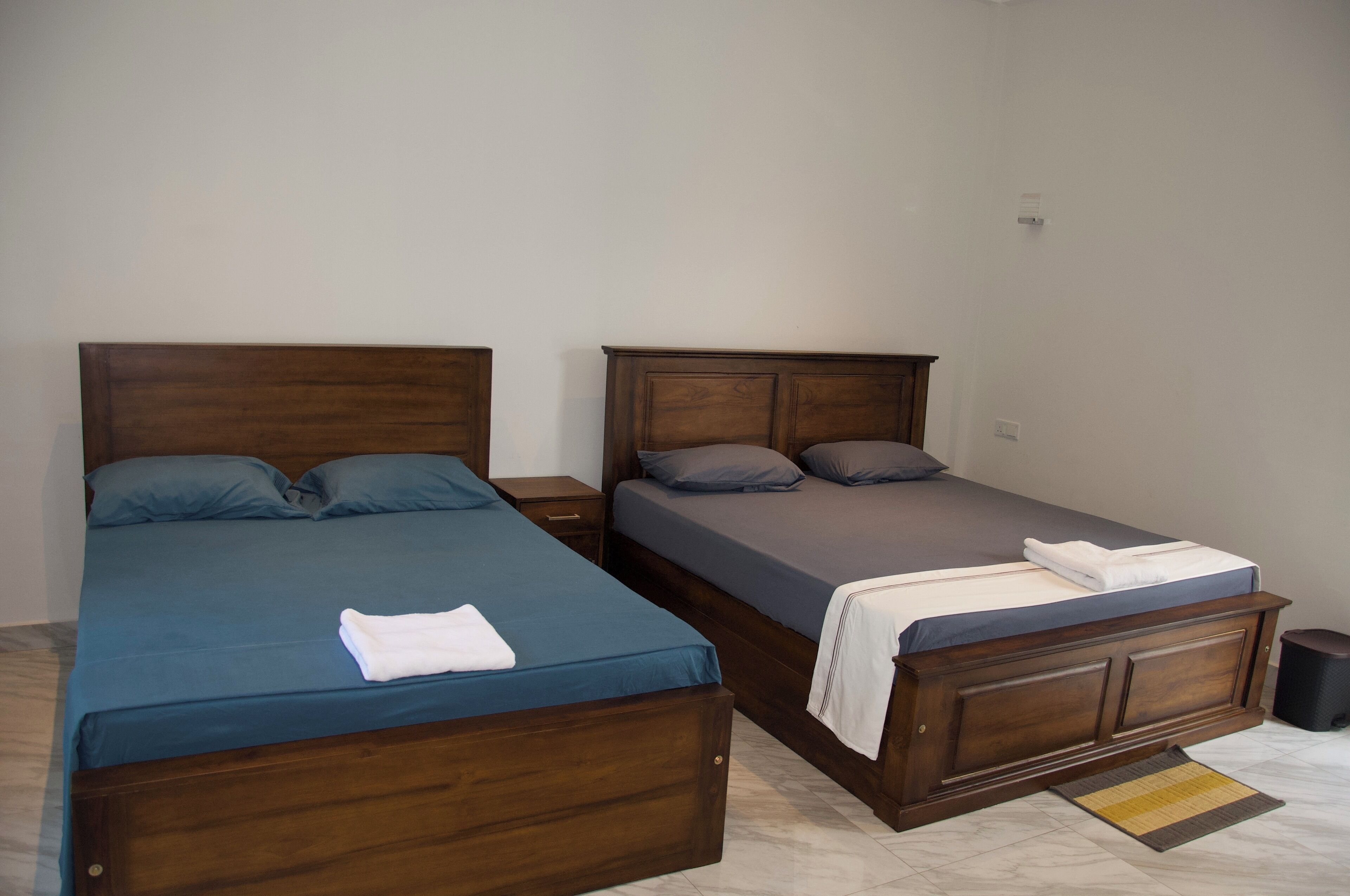 Dos camas de madera con ropa de cama azul y gris, una mesita de noche pequeña y un bote de basura.
