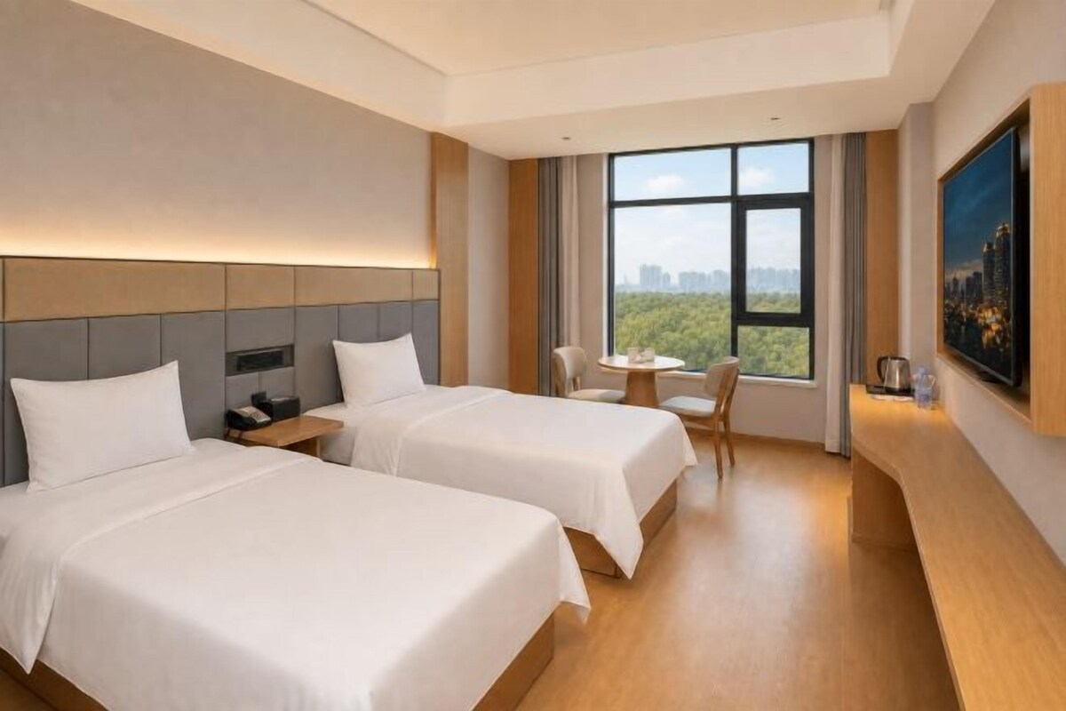 Habitación de hotel con dos camas, una mesita, una silla y un ventanal con vista a la ciudad.