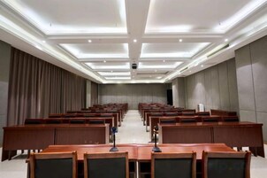 Un salón de conferencias con filas de mesas y sillas de madera, iluminación empotrada en el techo y una pantalla grande al frente.