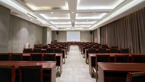 Une salle de conférence avec des rangées de tables et de chaises en bois, un écran de projection à l’avant et un plafond doté d’un éclairage encastré.