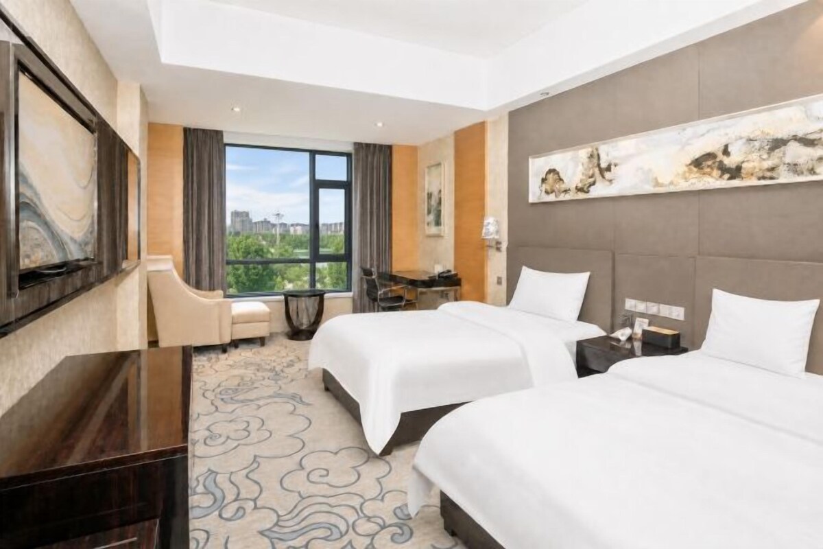 Habitación de hotel con dos camas, un escritorio, una silla, un televisor y un ventanal con vistas a la ciudad.