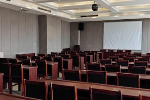 Un salón de conferencias con filas de mesas y sillas de madera, una pantalla grande y un micrófono en el techo.