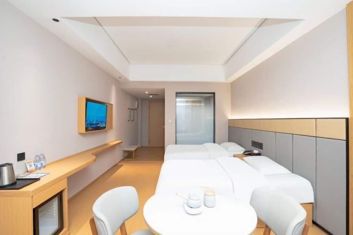 A modern hotel room with a long bed, a desk with a coffee maker, a television, and a glass door leading to a hallway.