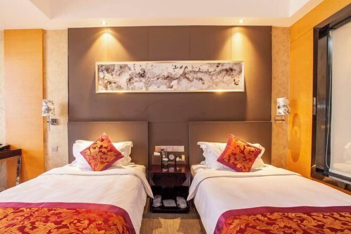 A hotel room with two beds, a nightstand, a painting above the beds, and a window on the right.