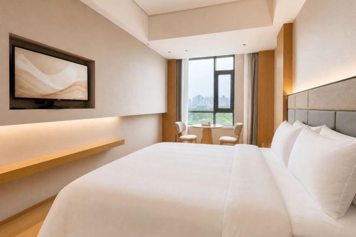 Habitación de hotel con una cama grande, televisión montada en la pared, una mesita con sillas y una ventana con vista a la ciudad.