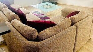 Een woonkamer met een grote L-bank, een glazen salontafel en een flatscreen televisie die aan de muur is bevestigd.