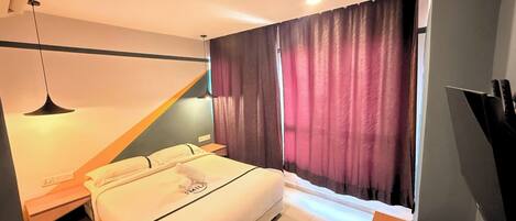 Habitación de hotel moderna con cama, mesitas de noche, un escritorio con computadora y control remoto, y una ventana con cortinas.