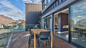 A modern outdoor dining area with a wooden deck, metal chairs, and a glass railing overlooking a mountainous landscape. - Stunning lake Views from Queenstown townhouse (Queenstown)