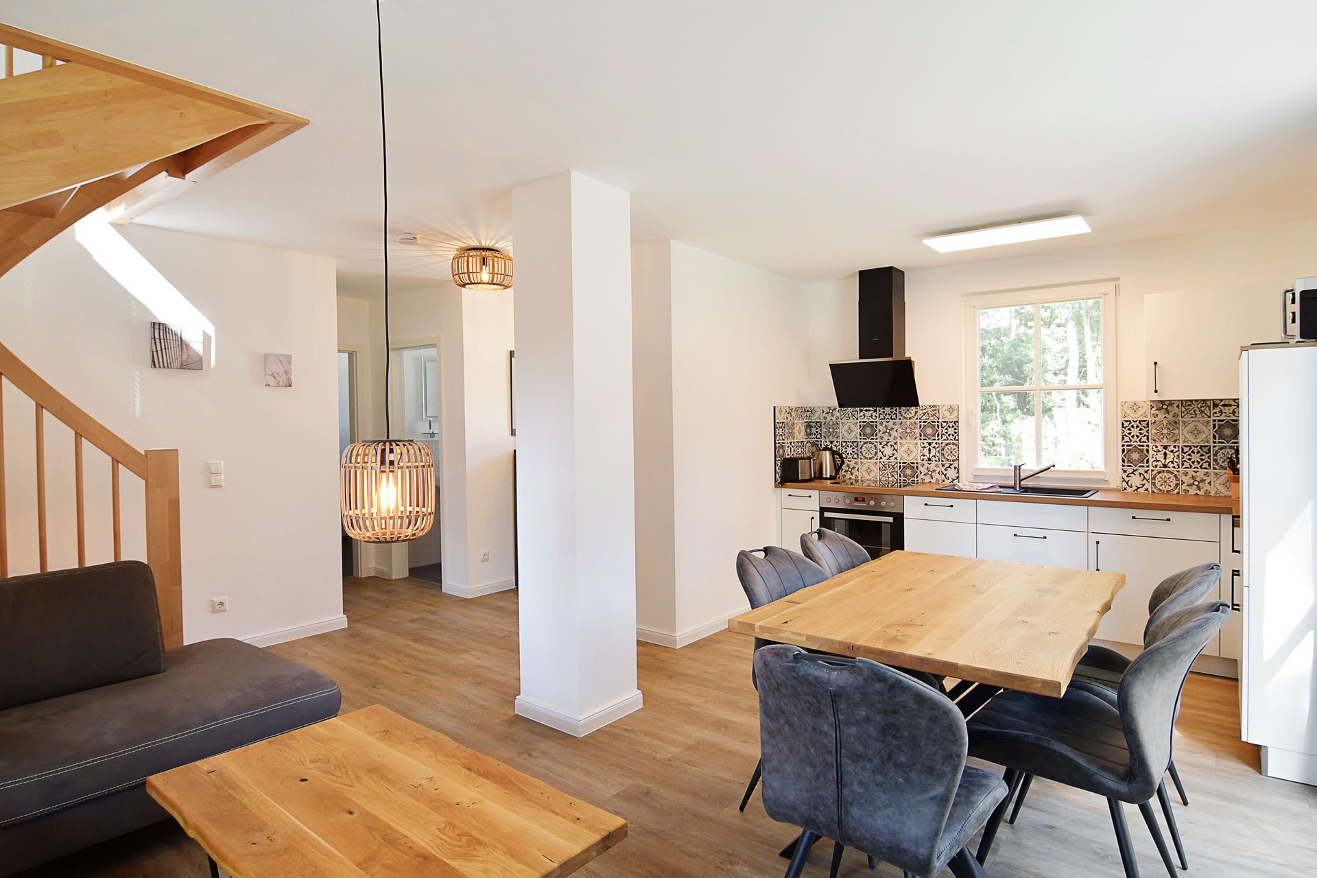 A modern kitchen with a wooden table and chairs, a built-in oven, and a window with a view of greenery.