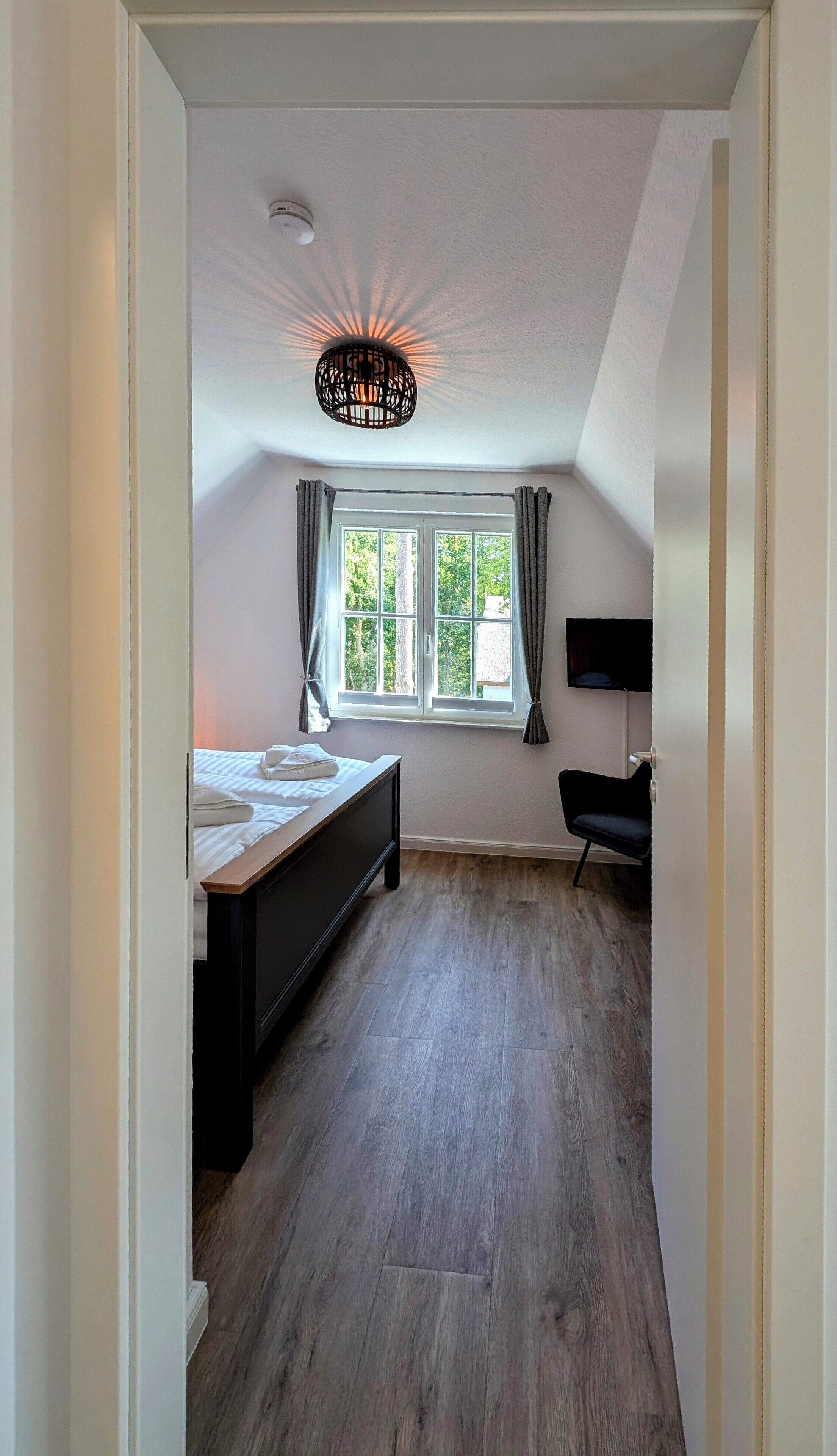 Et soveværelse med seng, skrivebord, stol, et vindue med gardiner og en loftlampe.