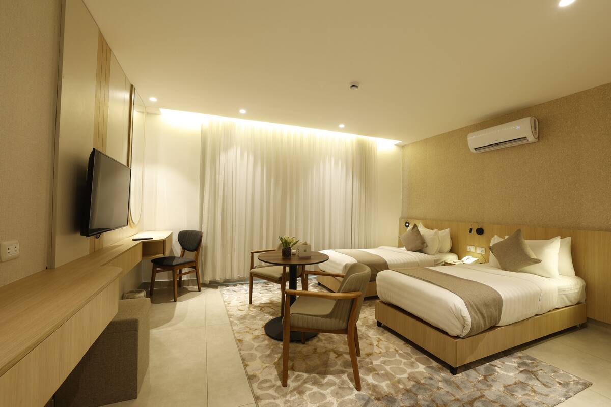 A modern hotel room with a flat-screen TV, two single beds, a desk with a chair, and a small table with a plant.