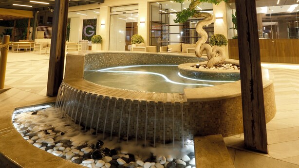 Un hotel con un llamativo logotipo 'G', un área de piscina ajardinada con rocas y una cascada, y una zona de estar al aire libre cubierta.