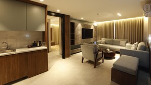 Un salón moderno con sofá, reposapiés y mesa de centro. Cuenta con una zona de cocina con lavabo y armarios. La habitación tiene un gran ventanal con cortinas.
