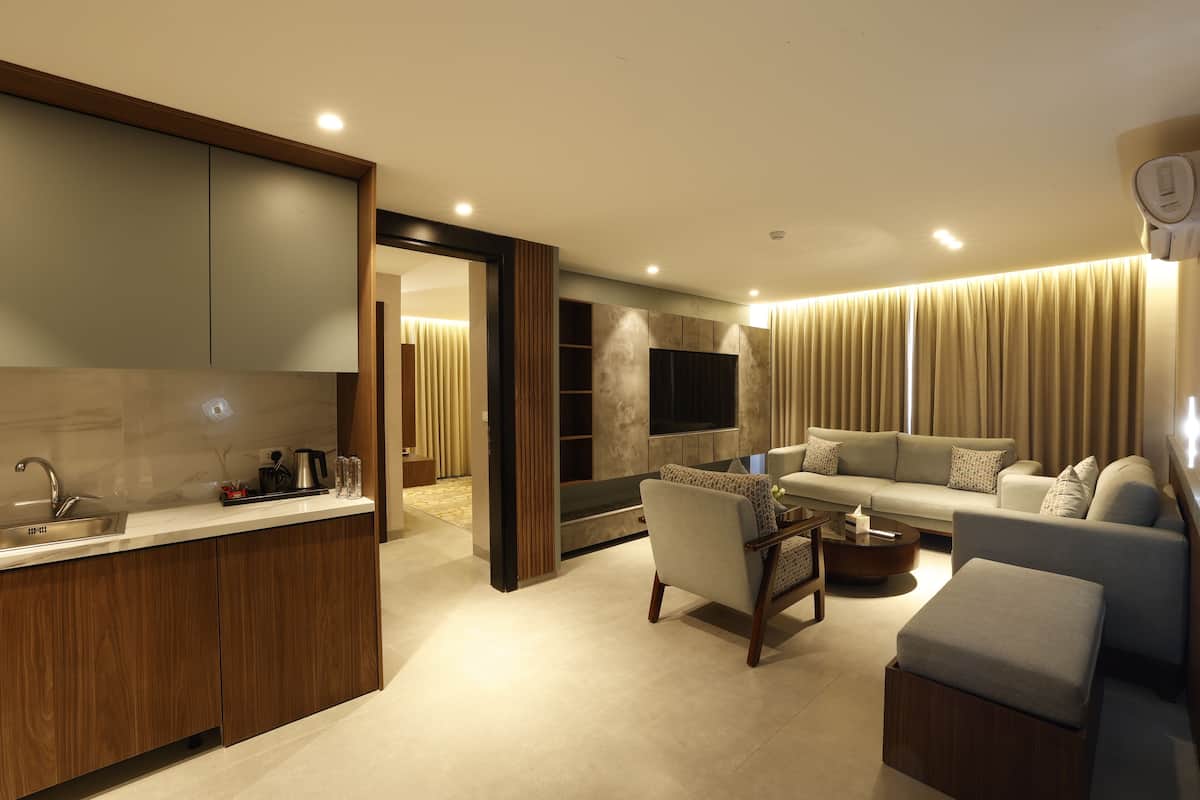 A modern living room with a sofa, ottoman, and coffee table. There is a kitchen area with a sink and cabinets. The room has a large window with curtains.