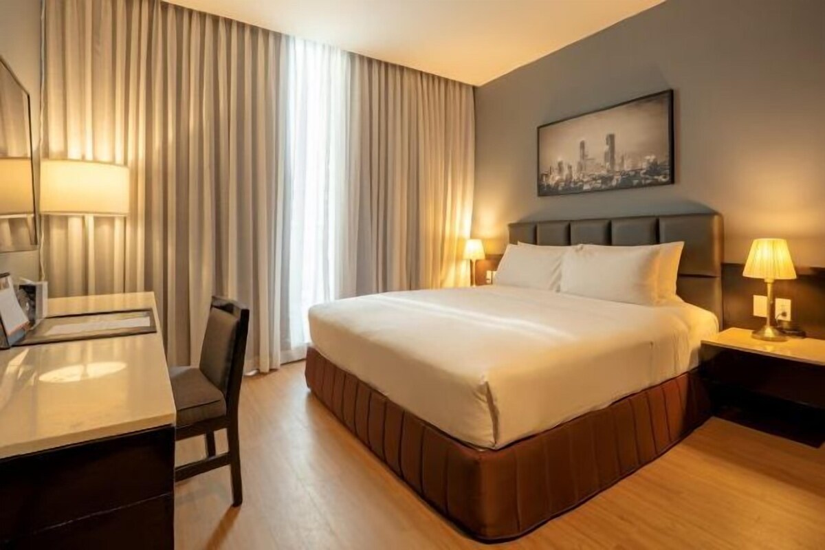 A hotel room with a large bed, a desk, a chair, and a nightstand with lamps.