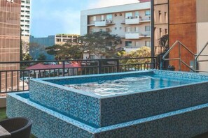 A rectangular hot tub with a tiled exterior, surrounded by a metal railing, set against a backdrop of residential buildings and a clear sky. - B Hotel Alabang (Muntinlupa)