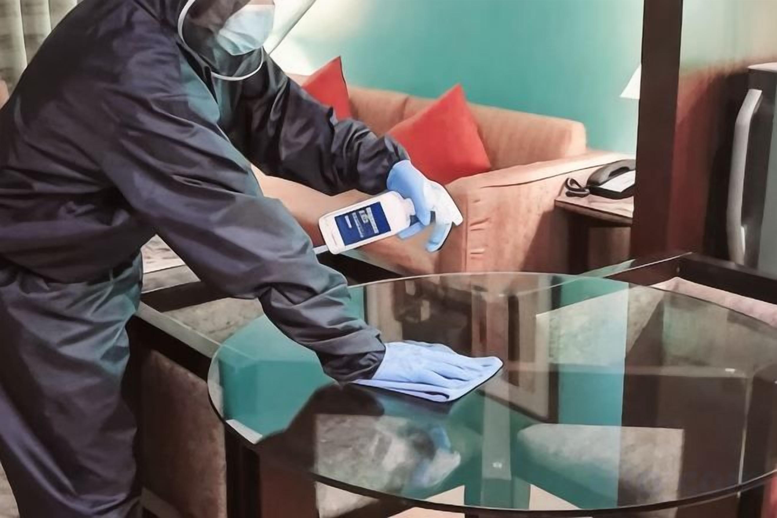 A person in protective gear cleaning a glass table in a room with a sofa and a telephone.