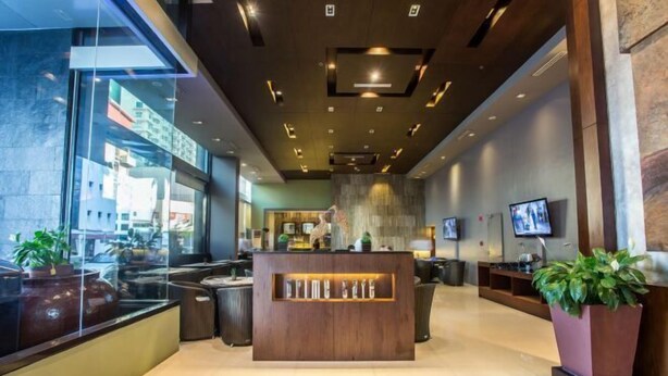 A modern reception area with a wooden reception desk, seating, and a large window offering a view of the city. - B Hotel Alabang (Muntinlupa)