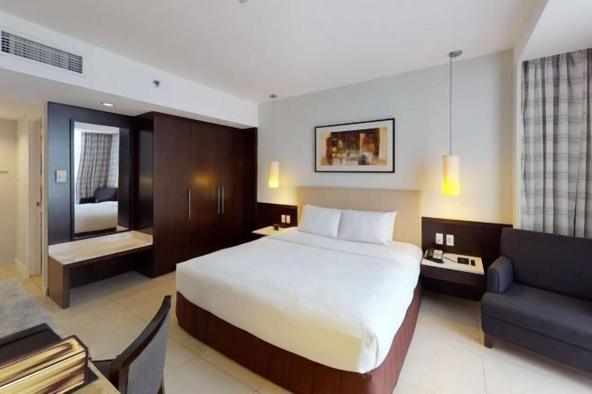 Ein modernes Hotelzimmer mit einem großen Bett, einem schrankhohen Holzschrank, Nachttischen mit Lampen, einem grauen Sofa und einem Gemälde an der Wand.
