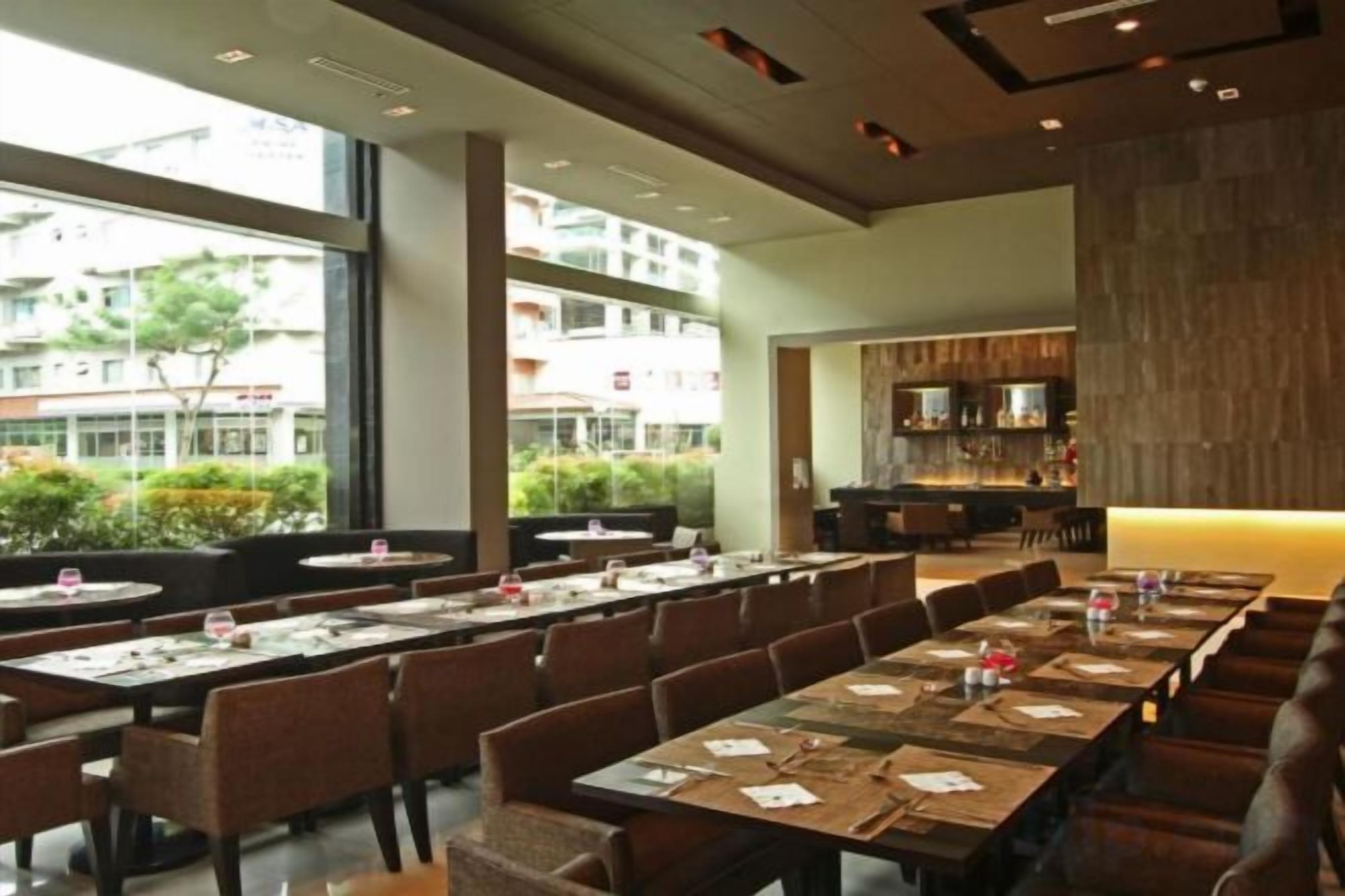 A restaurant with long tables and chairs, large windows, and a kitchen area.