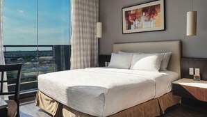 A hotel room with a large bed, a desk, a chair, a view of the city, and a painting on the wall. - B Hotel Alabang (Muntinlupa)
