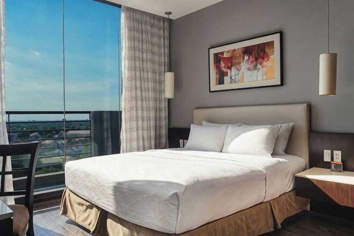 Ein Hotelzimmer mit einem großen Bett, einem Schreibtisch, einem Sessel und Blick auf die Stadt, sowie einem Bild an der Wand.