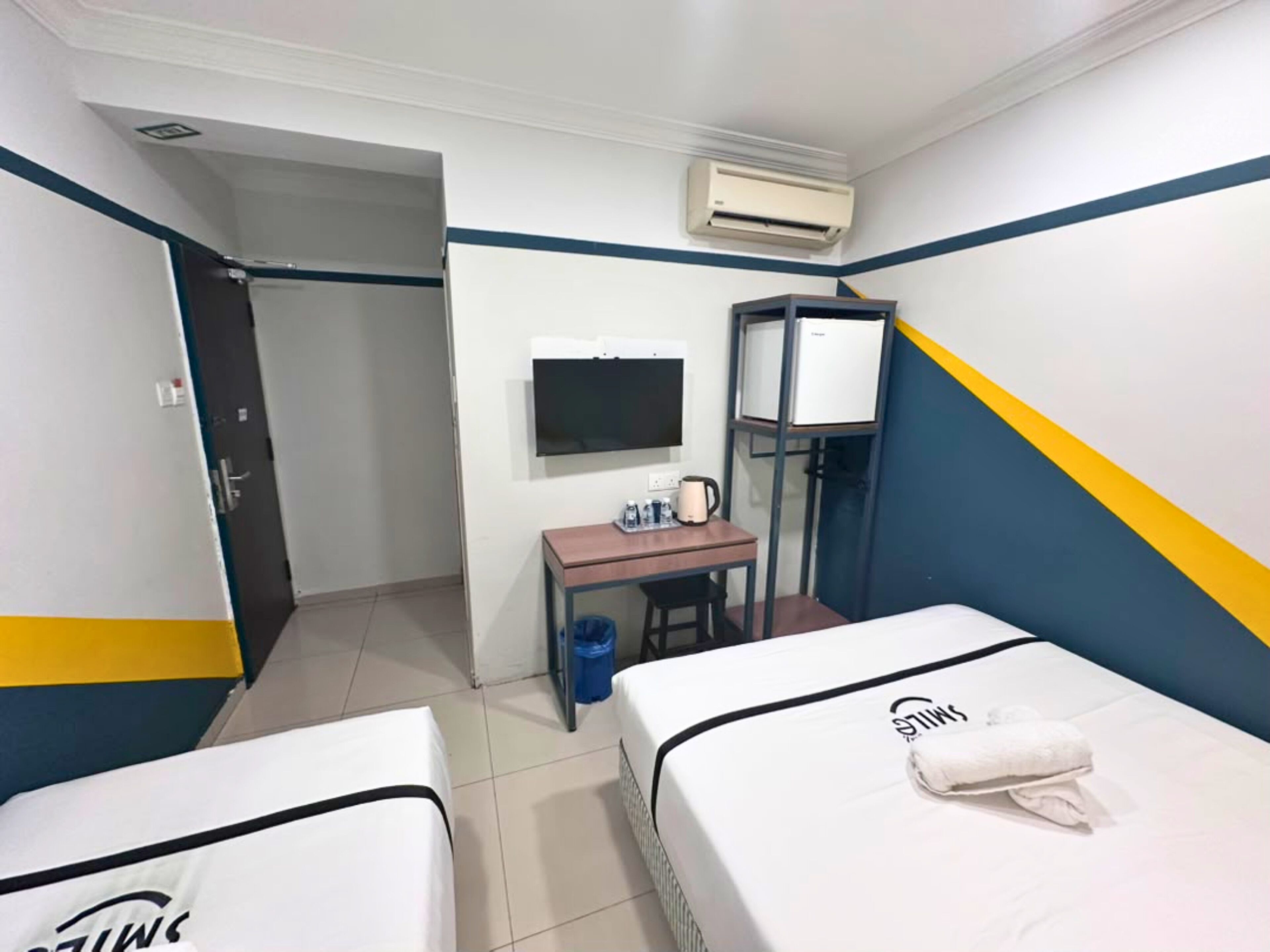 A hotel room with two beds, a desk, a television, an air conditioner, and a wardrobe.