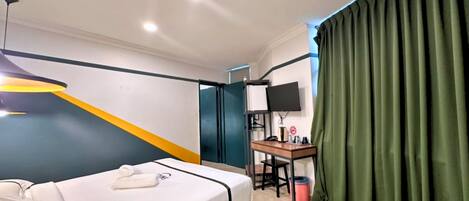 A hotel room with a bed, a desk with a computer, a television, and a green curtain.