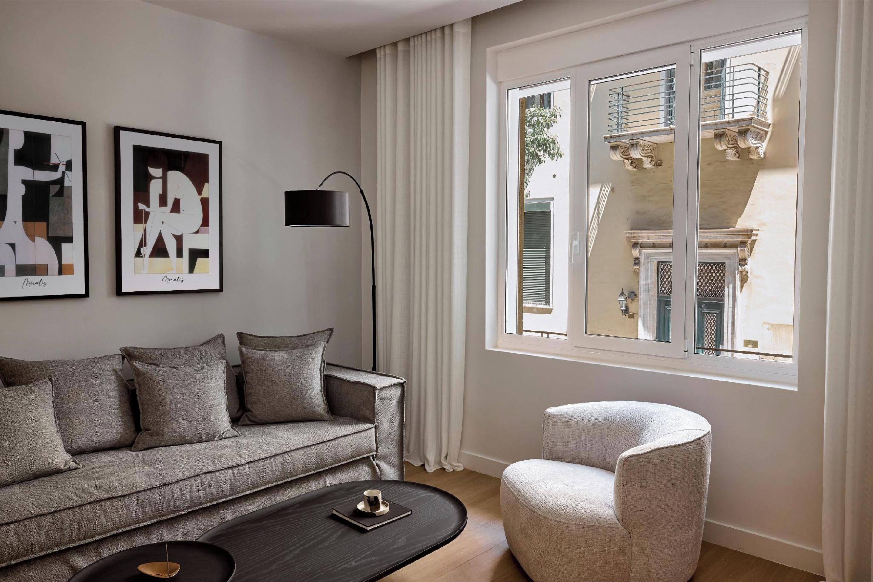 A modern living room with a grey sofa, a side table, a coffee table, and a chair. There are two framed artworks on the wall and a large window with a view of a balcony.