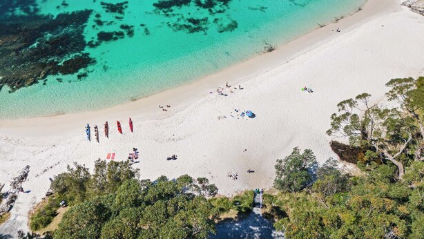 Vista aérea de una playa con aguas turquesas cristalinas, arena blanca y vegetación escasa.