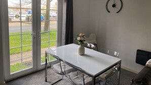 Una habitación moderna con una mesa blanca, sillas transparentes, un reloj en la pared y vistas a barcos a través de puertas correderas de vidrio.