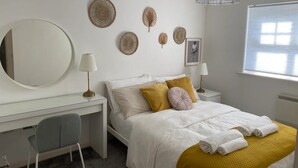 Un dormitorio moderno con un escritorio blanco, una cama con ropa de cama blanca y un plaid color mostaza, un espejo redondo y decoración en la pared.