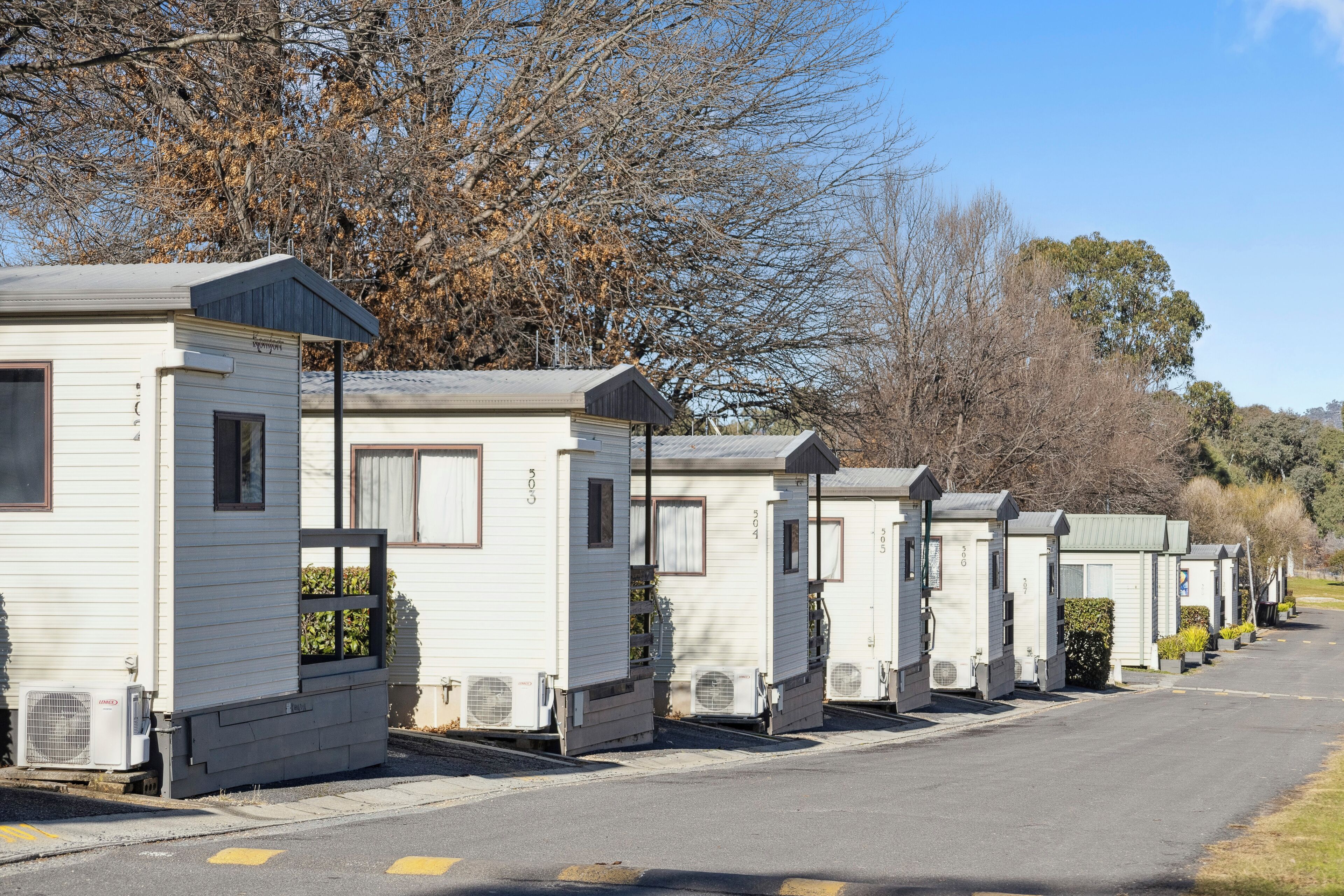 A row of white mobile homes with air conditioning units, a paved road, and trees in the background.