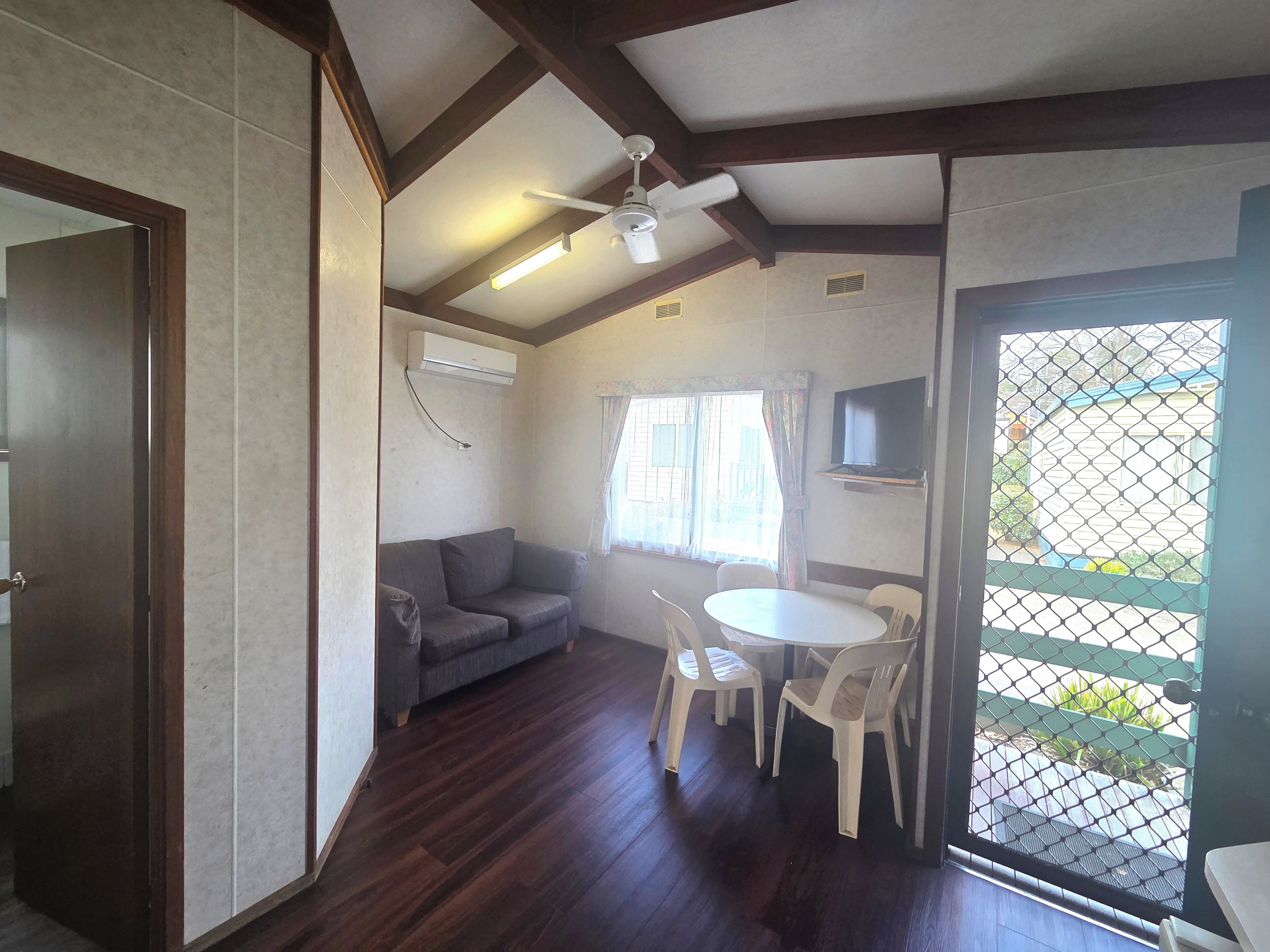 A compact room with a ceiling fan, a sofa, a small table with chairs, and a TV mounted on the wall.