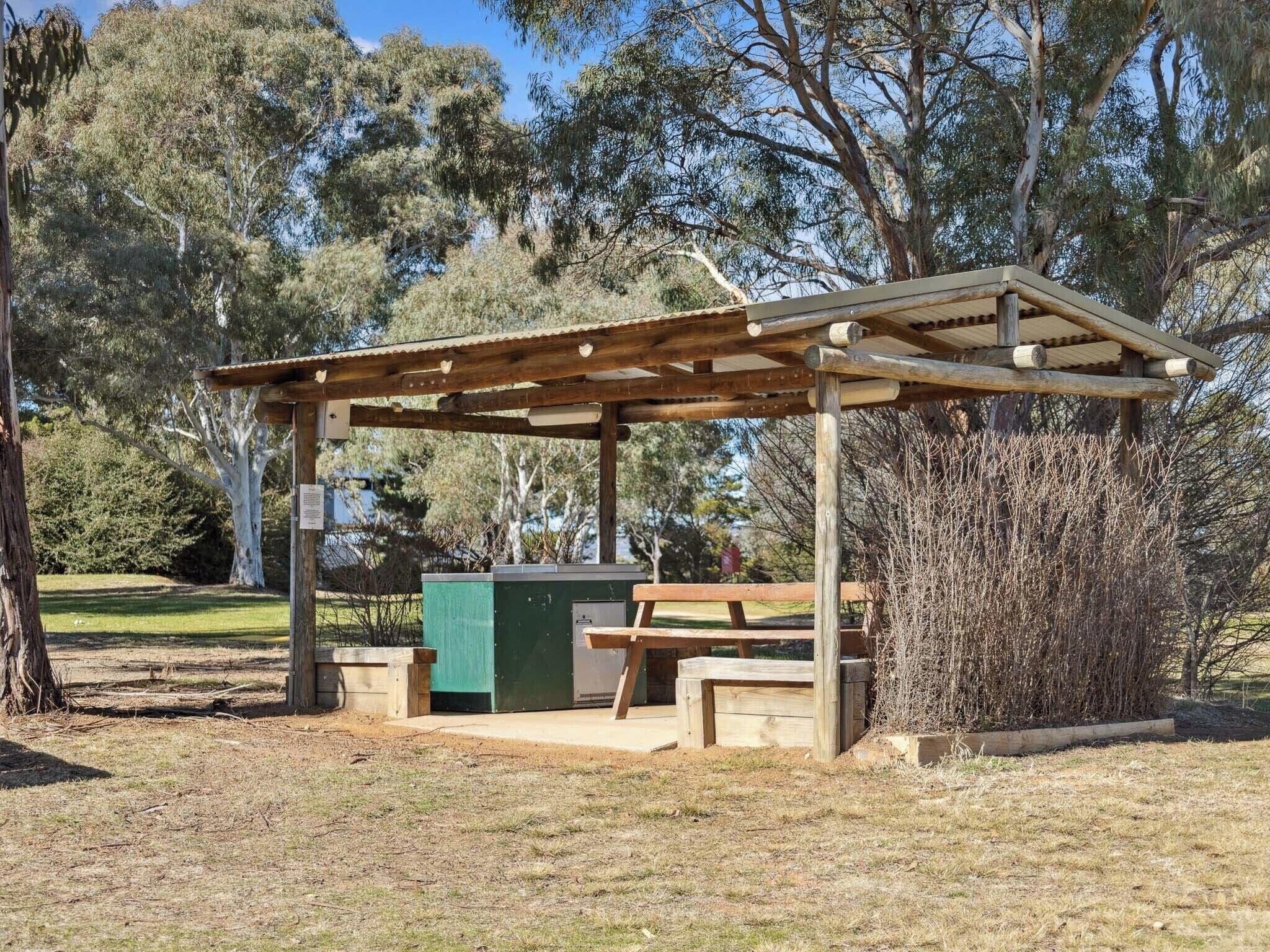 Une pergola en bois, agrémentée de bancs et d’une table, entourée d’arbres et d’une pelouse.