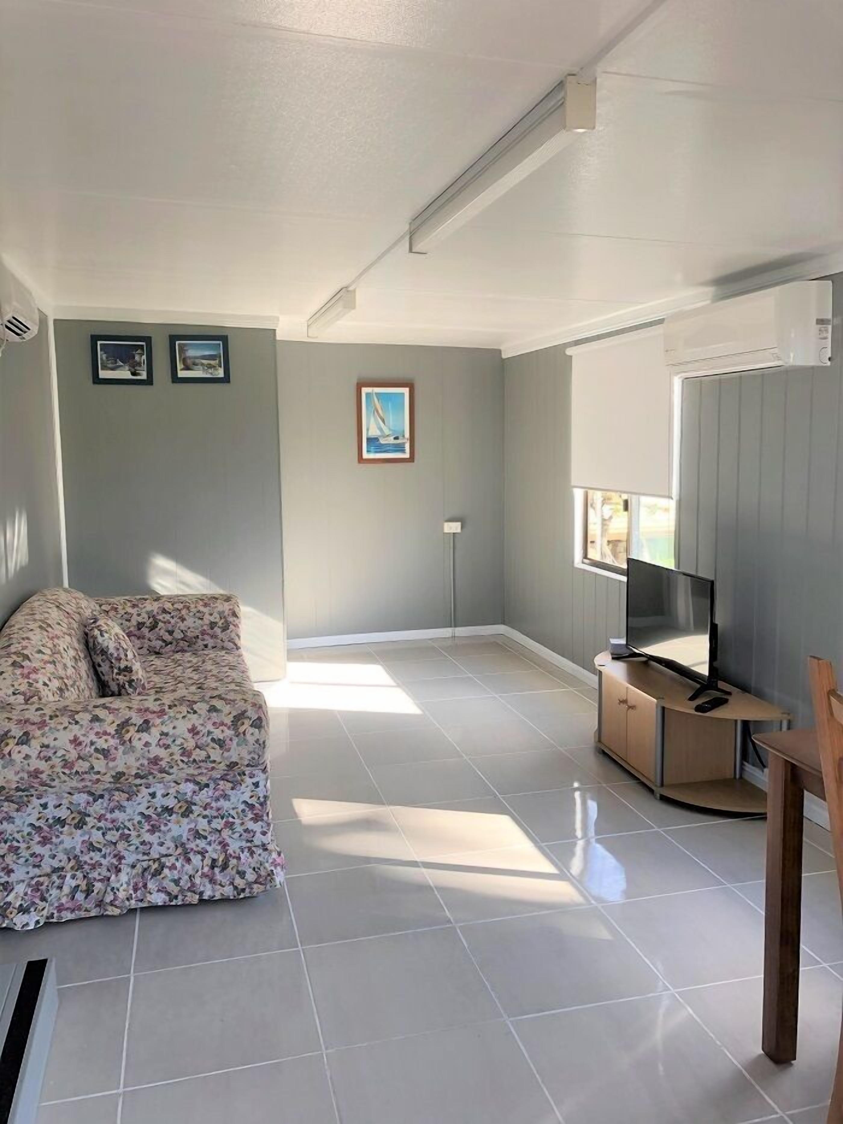A spacious living room with a floral sofa, a flat-screen TV on a wooden stand, and a large window allowing natural light.