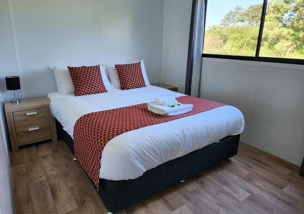 A neatly made bed with a red and white patterned bedspread, a white pillow, and a red pillow. There is a wooden nightstand with a lamp and a drawer to the left of the bed. The room has a large window on the right side, providing a view of greenery outside.