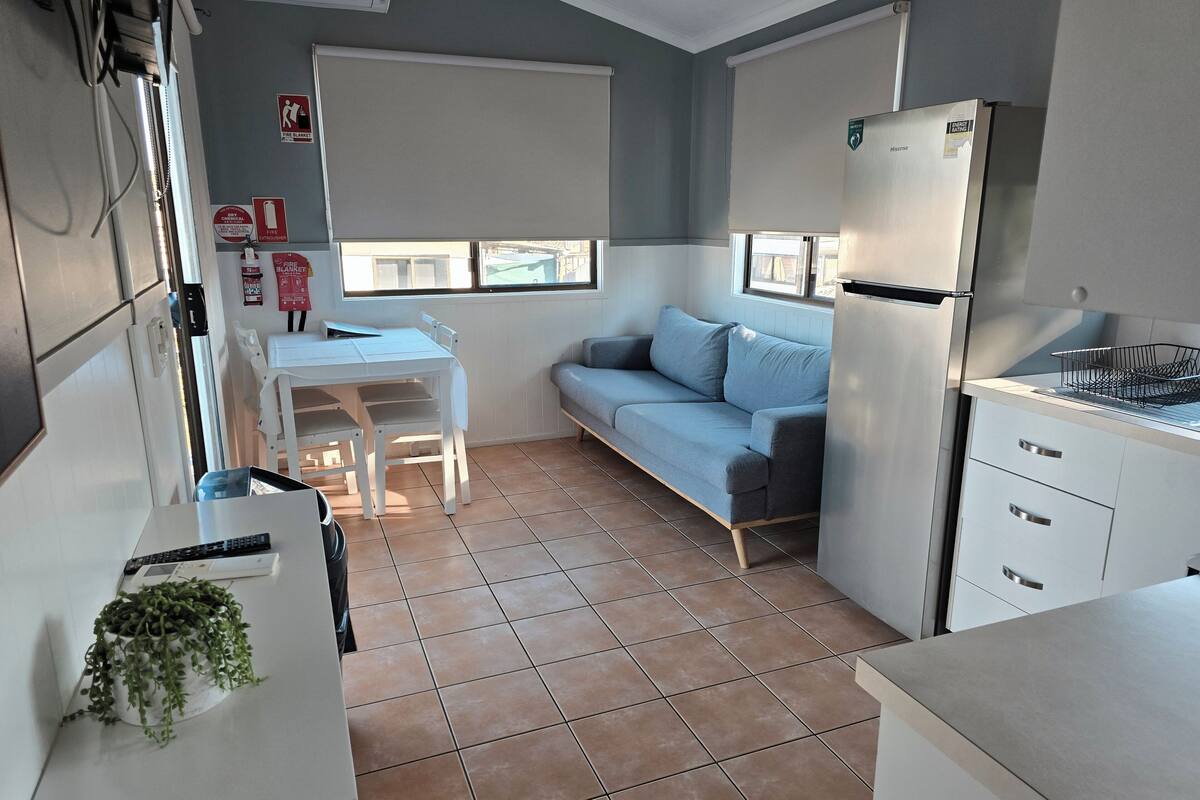 A compact kitchen with a refrigerator, a dining table with chairs, a sofa, and a small countertop with a sink.