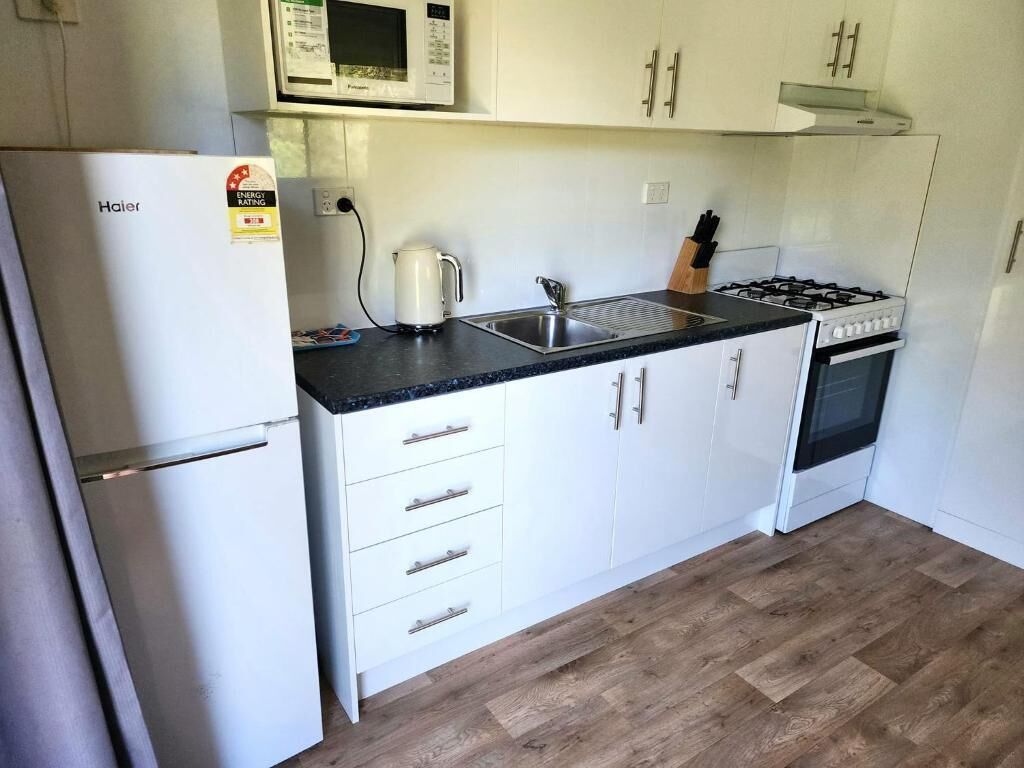 Ett kompakt kök med vita skåp, en diskho i rostfritt stål, en gasspis och ett kylskåp från Haier.