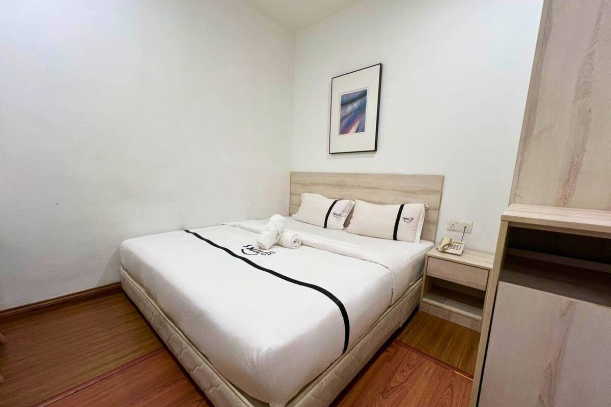 Een opgemaakt bed met een witte deken en zwarte rand, een nachtkastje met een telefoon, en een lijst met een foto aan de muur.