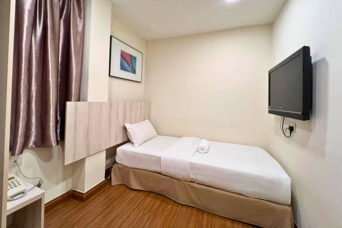 Een kleine kamer met een enkel bed, een houten vloer, een nachtkastje met een telefoon, een wandtelevisie en een ingelijst schilderij.