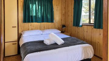 A single bed with a dark headboard, white bedding, and a folded towel on the bed. There is a nightstand with a lamp, and the room has wooden walls and a window with curtains.