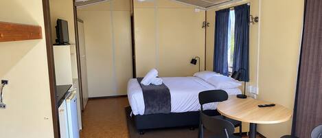 Habitación pequeña con cama, una mesita, sillas, televisor y ventana con cortinas.