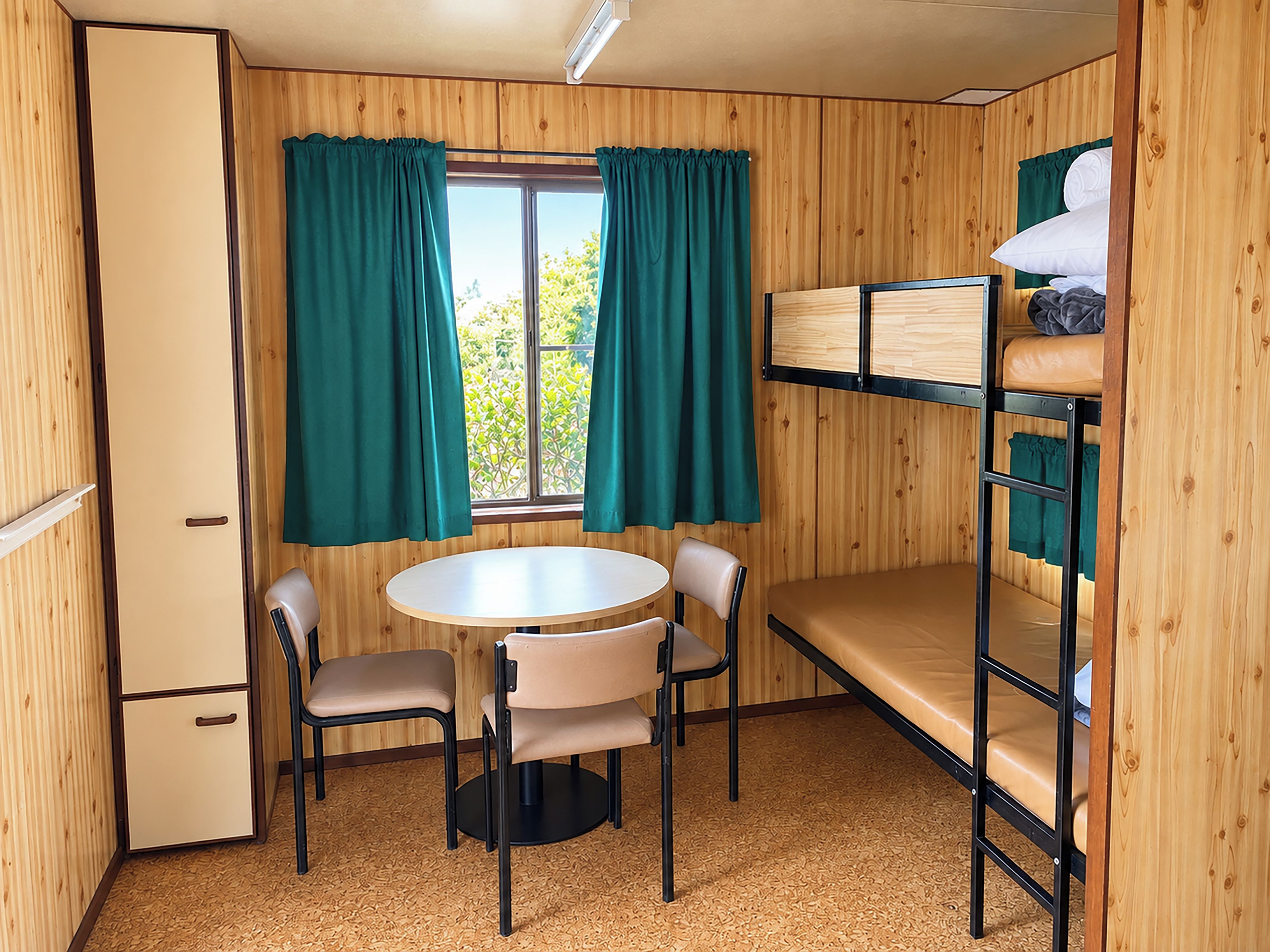 A compact room with a bunk bed, a small kitchenette, a round table with two chairs, and a window with curtains.